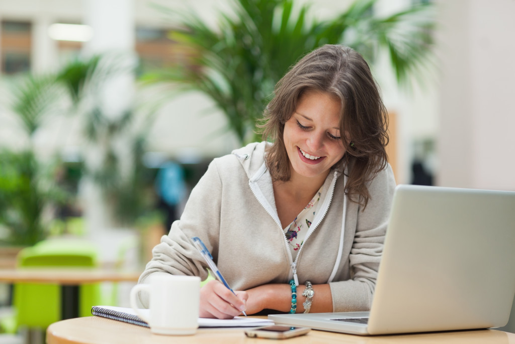 Smiling female student doing homework by laptop at cafeteria table